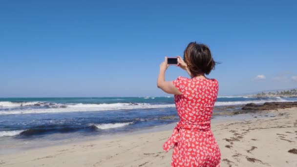 Mujer atractiva joven sosteniendo el teléfono de la cámara y tomando fotos de las olas de mar tormenta de pie en la playa de Cyprus. Movimiento lento — Vídeos de Stock