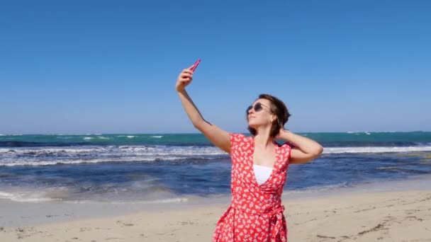 Young beautiful woman in red dress taking selfie with her camera phone on the sea beach with strong wind and waves. — Stock Video