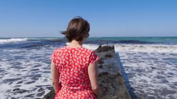 Νεαρή ελκυστική λευκή γυναίκα που περπατάει στην προβλήτα φορώντας κόκκινο φόρεμα. Ξυπόλητα πόδια περπατούν προς τη θάλασσα στην προβλήτα. — Αρχείο Βίντεο