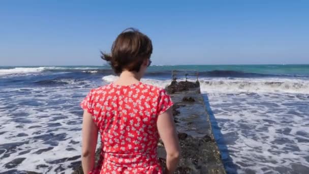 Jonge aantrekkelijke blanke vrouw die over de pier loopt in een rode jurk. Barefoot benen lopen naar de zee op de pier. Langzame beweging — Stockvideo