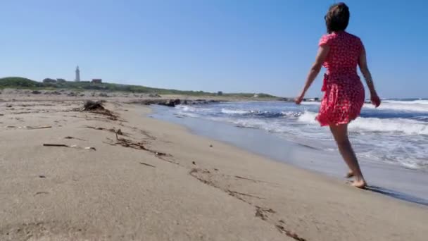 Boş plaj boyunca koşan kırmızı elbiseli genç bir kadın. Güçlü dalgalar sıçrıyor ve rüzgar esiyor. Arka planda deniz feneri — Stok video