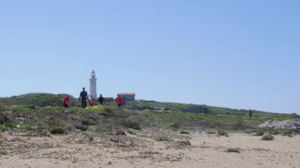 Les gens marchant le long du chemin de plage près de la mer avec phare sur le fond à Paphos, Chypre — Video