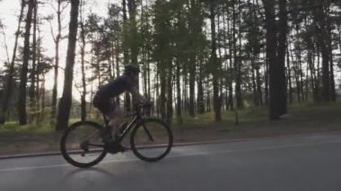 Genç ve güzel kız bisikletçi rolde bisiklet sürüyor. Arka planda ağaçların arasından güneşli bir çekim. Bisiklet konsepti. Yavaş çekim