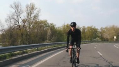 Gün batımında kameraya doğru giden bir yol bisikleti üzerinde Odaklanmış Bisikletçi. Siyah mayo ve şort giyen bisikletçi. Bisiklet konsepti.