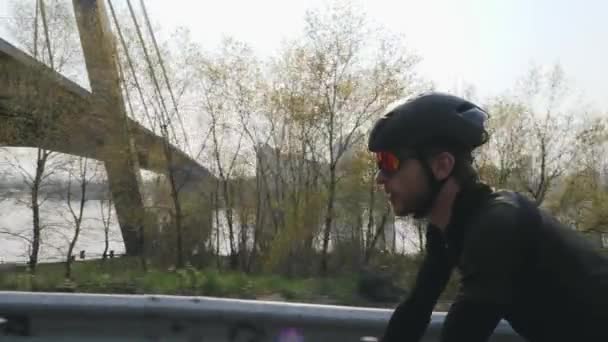 Concentrato ciclista fiducioso su una bicicletta. Il sole splende. Fiume e ponte sullo sfondo. Vista laterale da vicino. Concetto bicicletta . — Video Stock