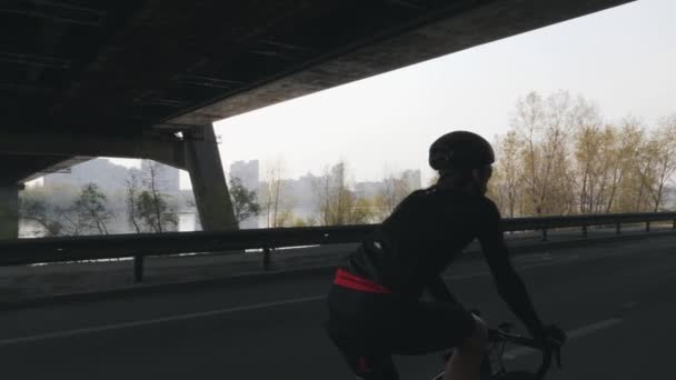 Ajuste y ciclista fuerte pedaleando una bicicleta con ciudad, puente y río en el fondo. Concepto de ciclismo y entrenamiento. Movimiento lento — Vídeos de Stock