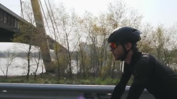 Focused confident cyclist on a bicycle. Sun shining through. River and bridge on background. Close up side view. Cycling concept. Slow motion — Stock Video