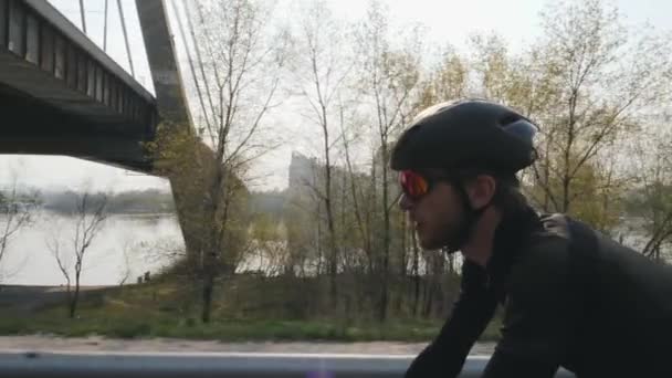 Primer plano de ciclista en bicicleta. Ciclista barbudo con casco negro y gafas de sol montando bicicleta con puente y río en el fondo. Movimiento lento — Vídeo de stock