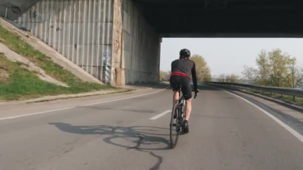Молодой спортсмен катается на велосипеде под мостом. Велоспорт тренировки для гонки. Мужчина-велосипедист в черном костюме и шлеме . — стоковое видео