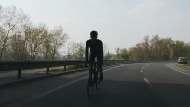 Ciclista fuerte y en forma con barba con traje negro, casco y gafas de sol montando en bicicleta fuera de la silla. Disparo de seguimiento frontal. Movimiento lento — Vídeos de Stock