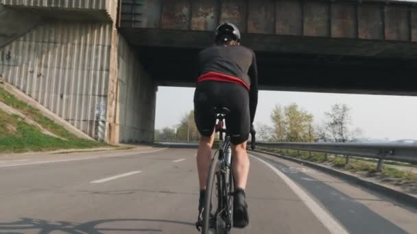 Männlicher Radfahrer auf einem Fahrrad. zurück folgen Schuss. Radfahrer mit schwarz-rotem Outfit, Helm und Brille. kräftige Beinmuskeln. Zeitlupe — Stockvideo