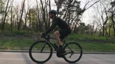 Bisikletçi parkta siyah bir profesyonel yol bisikleti sürme siyah mayo ve şort giyiyor. Yan takip atış. Bisiklet konsepti. Yavaş çekim