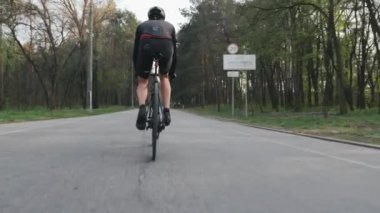 Bisikletle pedal çeviren bisikletçi bacak kası görünümüne yakın. Parktaki bisiklet sürücüsü siyah spor kıyafetler giyiyor. Yavaş çekim