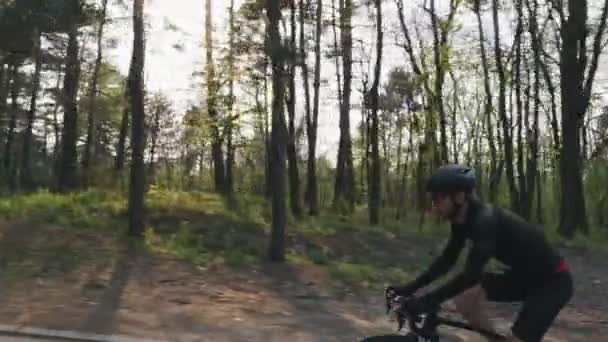 Fiatal kerékpáros szakállas fekete ruhában, sisakkal és napszemüveggel biciklizik a parkban. A nap átsüt a fákon. Kerékpáros koncepció. Lassú mozgás. — Stock videók