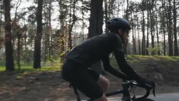 黒いジャージ、ショートパンツ、ヘルメット、サングラスを着用したサイクリストペダリングバイクのクローズアップショット。公園内のバックロードカーボン自転車。スローモーション — ストック動画