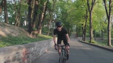 Odaklanmış güçlü uygun bisikletçi yokuş yukarı bizim eyer siyah spor giyim, gözlük ve kask giyiyor sürme. Profesyonel bisikletçi kavramı. Yavaş çekim