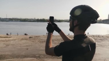 Bisikletçi tepede duran nehir ve şehir fotoğraflarını çekmek kameralı telefon tutan. Köprü ve güneş arka planda parlıyor. Yavaş çekim