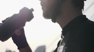Siyah kıyafet, kask ve güneş gözlüğü giyen siyah şişeden odaklanmış sakallı bisikletçi içme suyu yakın portresi.