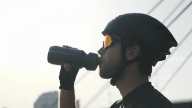 Kırmızı sakallı kendine güvenen bisikletçi, kask ve güneş gözlüğü takan şişeden içme suyu içiyor. Arka planda köprü ve nehir. Yavaş çekim