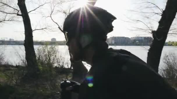 Vista de cerca del ciclista con barba bebiendo agua mientras monta en bicicleta. El sol brilla a través de los árboles reflejando en el casco y las gafas de sol . — Vídeos de Stock