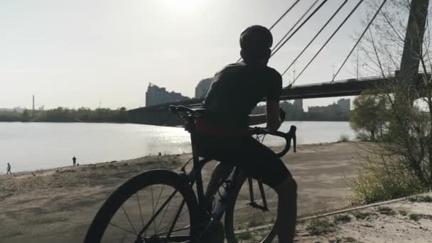 Kräftiger, athletischer Radfahrer, der auf dem Fahrrad sitzt und weit voraus blickt. Radfahrer träumt von der Zukunft. Brücke und Fluss im Hintergrund. — Stockvideo