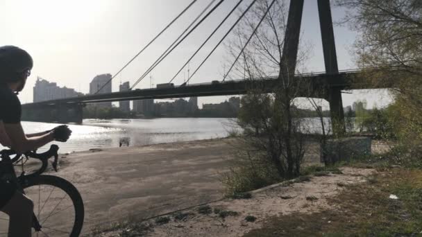 Cycliste en noir vêtements de sport sur vélo noir assis et regardant vers la rivière et le pont rêvant de l'avenir. Concept de vélo. Mouvement lent — Video