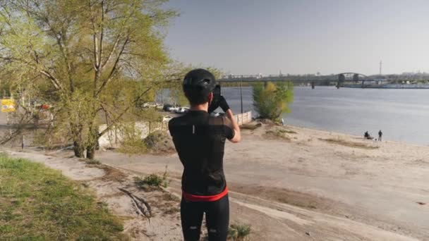 Ciclista con traje negro, casco y gafas de sol sostiene su cámara del teléfono celular y toma fotos del río de la ciudad. Deportista masculino con cámara telefónica. Movimiento lento — Vídeo de stock