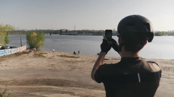 Male cyclist holding phone camera and taking pictures of river, bridge and city. Bearded cyclist making photos with his phone camera in hands. Slow motion — Stock Video
