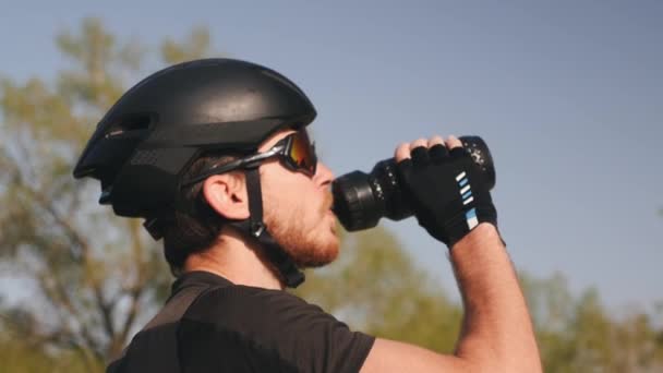 Close up portrait of triathlete in black helmet and glasses drinking water. Bearded male cyclist drinks water from water bottle. — Stock Video