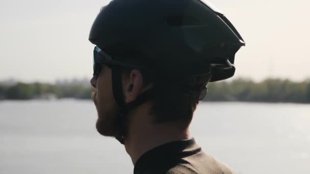 Cycliste barbu portant un casque noir et des lunettes de soleil regardant vers l'horizon. Rivière et pont en arrière-plan. Concept de vélo . — Video