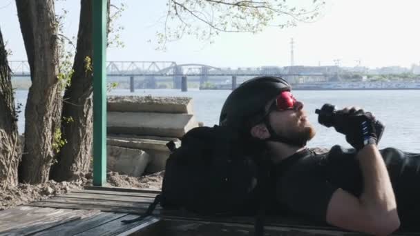 Rowerzysta z brodą wody napoje z bidon leżąc na ławce odpoczynku po treningu. Miejskie miasto i rzeka na tle. — Wideo stockowe