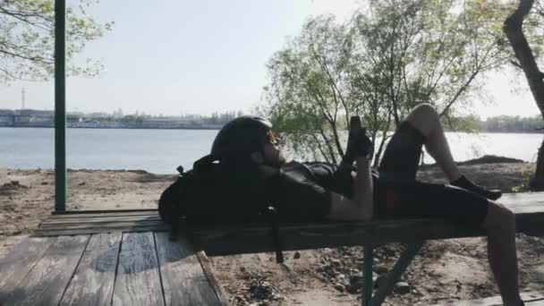 Der schlanke kräftige Radfahrer ruht sich auf der Bank aus, das Handy in der Hand klickt und scrollt auf dem Bildschirm. Fluss und Stadt im Hintergrund. Sonnenuntergang in der Stadt. — Stockvideo