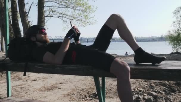 Ciclista com barba vermelha deitada no banco com mensagens de celular e alimentação de rolagem. Ciclista esportivo segurando telefone enquanto descansa no banco . — Vídeo de Stock