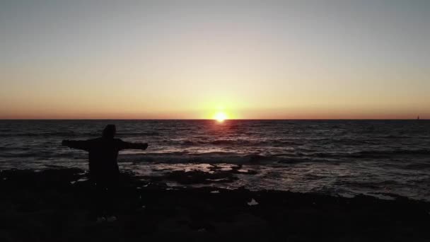 岩の多いビーチに立つ若い女性のシルエットは、太陽の道と波状の海の上に夕日の両側に広がる手で。空中ドローンビュー — ストック動画
