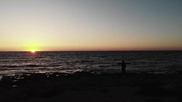 Černá temná silueta člověka stojící na pláži s otevřenou náručí, která hledí na západ slunce nad oceánem s malou lodí na obzoru. Vzdušný výhled — Stock video
