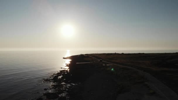 Εναέρια βολή του ήλιου λάμπει ενάντια στην κάμερα δημιουργώντας δρόμο ήλιου με θάλασσα και παραλία με πεζούς που περπατούν μόνες ακτογραμμή. — Αρχείο Βίντεο