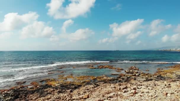 Tiro aéreo de ondas do oceano atingindo praia rochosa. Céu azul com nuvens brancas no horizonte. Dia ventoso ensolarado em Chipre Paphos — Vídeo de Stock