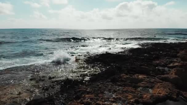 Disparo aéreo sobre las olas del mar golpeando la playa rocosa salpicando contra las rocas. Cielo azul con la carretera del sol en el horizonte. Océano en un día ventoso . — Vídeos de Stock