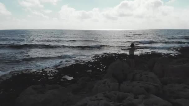 Dark silhouette of female person with arms wide open in air standing on cliff with strong waves hitting ocean beach. Aerial drone view — Stock Video