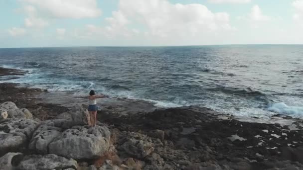 Woman silhouette with hands widespread in air on rocky cliff along ocean with waves hitting coastline. Female with arms wide open freedom standing on rock. Aerial shot. — Stock Video