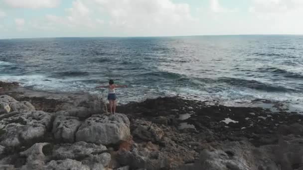 Young caucasian woman standing on big rock with arms wide open looking at stormy ocean sea with waves hitting seashore. Aerial drone fly over shot. — 비디오