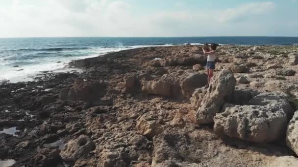 Mujer joven en las rocas tomando fotos con la cámara. Fuertes olas marinas golpeando la costa. Vista aérea del dron. Puesta de sol en cyprus — Vídeos de Stock