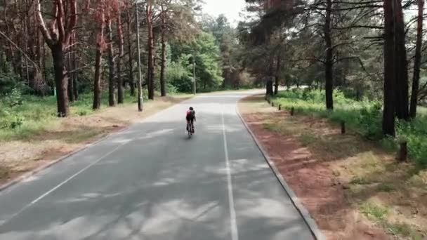 Bela menina esportiva jovem em uma camisa rosa está de bicicleta através do parque. Visão do drone traseiro . — Vídeo de Stock