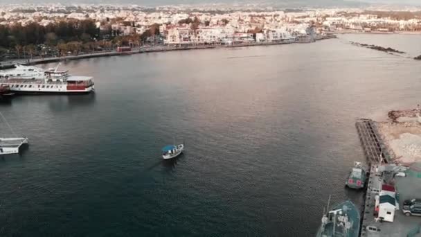 Drone view of little white fishing boat with blue roof sailing to open ocean from city harbor at sunset — Stock Video