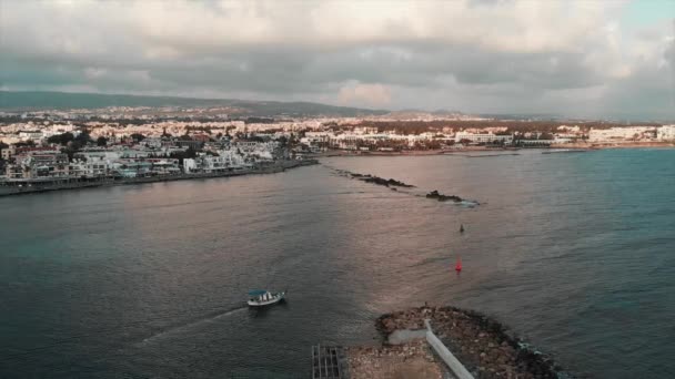 Small white fishing boat with blue roof sails into open sea at sunset with big resort city on background. Aerial drone view — Stock Video