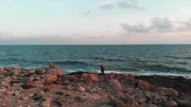 Genç bayan fotoğrafçı gün batımında büyük okyanus dalgaları olan güzel kayalık deniz kıyısında fotoğraf çekmek için bir yer arıyor. Genç ve çekici bayan turist kayalıklara çarpan dev deniz dalgalarının fotoğraflarını çekiyor.