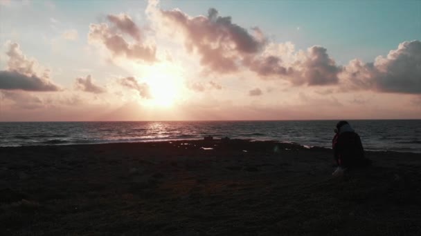 Jovem fotógrafa bonita sentada na costa rochosa e tirando fotos e vídeos do lindo pôr-do-sol rosa do oceano com sunroad e magnífico céu nublado em sua câmera profissional — Vídeo de Stock