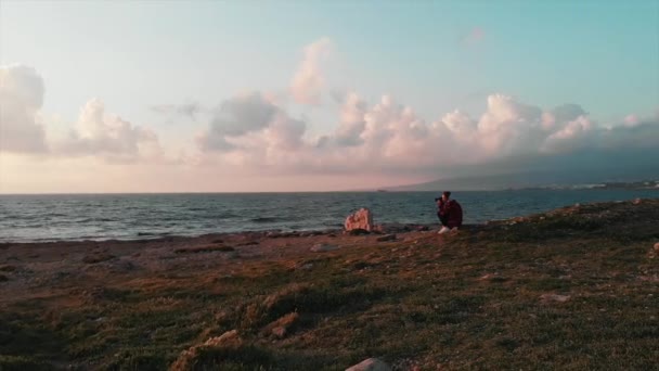 Молодая привлекательная женщина-фотограф сидит на скалистом пляже и фотографирует розовый красивый закат и великолепное облачное небо — стоковое видео