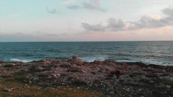 Gün batımında kayalık deniz kıyısından okyanus plajına giden kırmızı gömlekli çekici beyaz beyaz kadın gezgin — Stok video