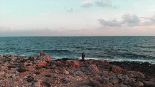 Jonge vrouwelijke fotograaf op zoek naar een plek om foto 's te maken aan prachtige rotsachtige kust met enorme oceaangolven bij zonsondergang. Jonge charmante vrouwelijke toerist neemt foto 's van enorme golven die tegen kliffen botsen — Stockvideo